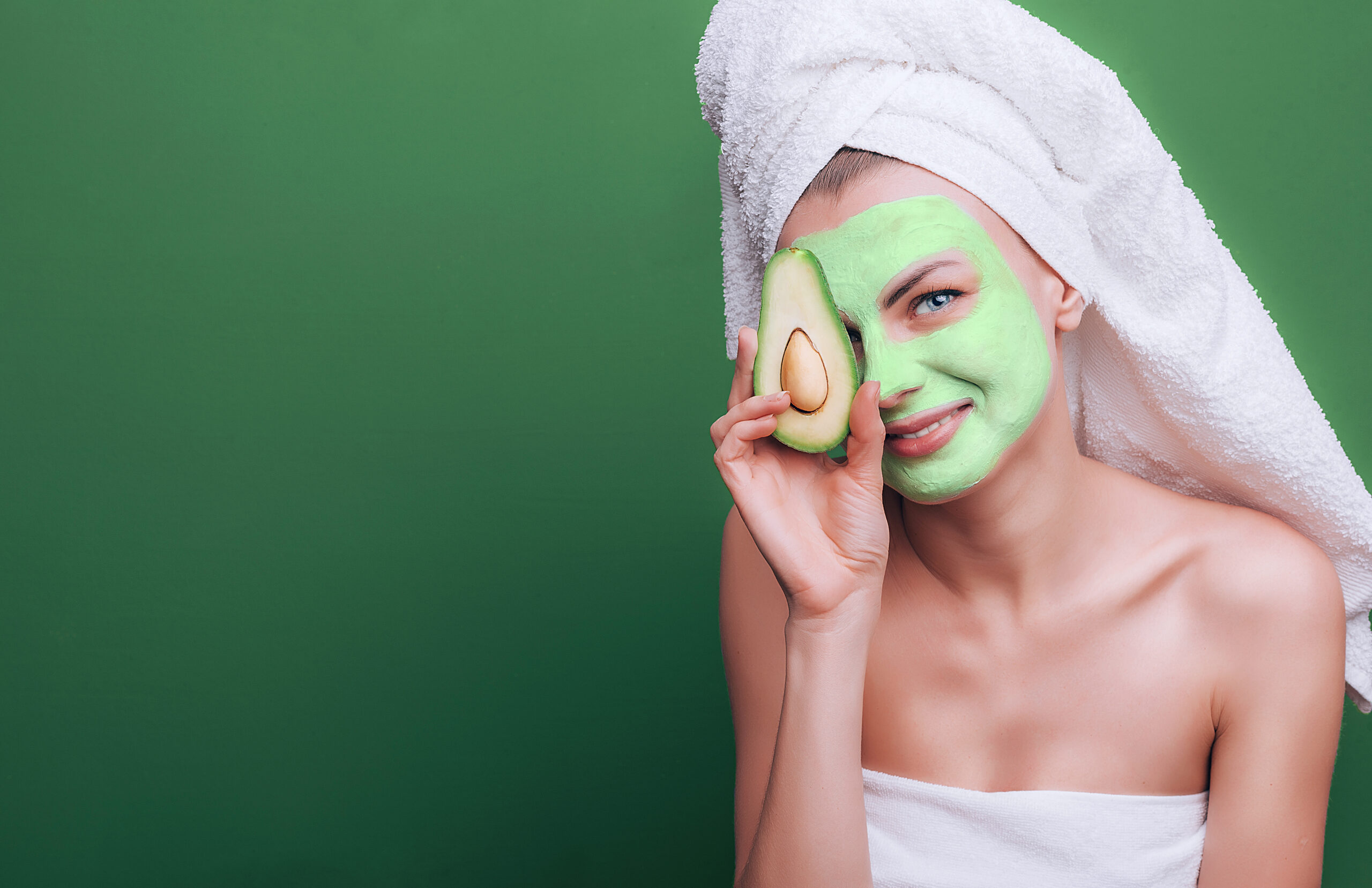 girl with a white towel on her head with a nutritious green mask on her face and an avocado in her hands on a green background with space for text